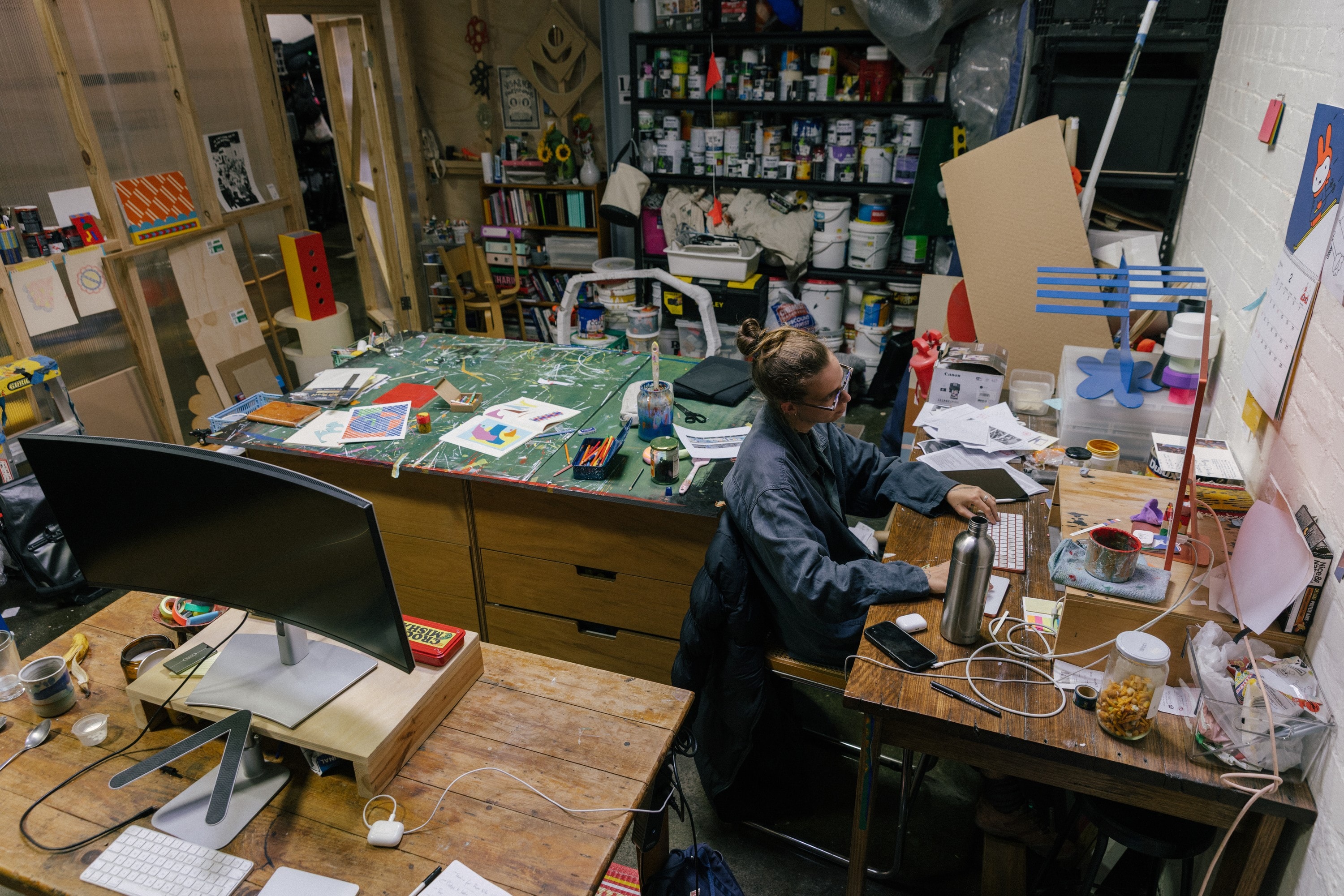 Abbey Rich working at a cluttered desk in the corner of the studio, shelves of paint behind