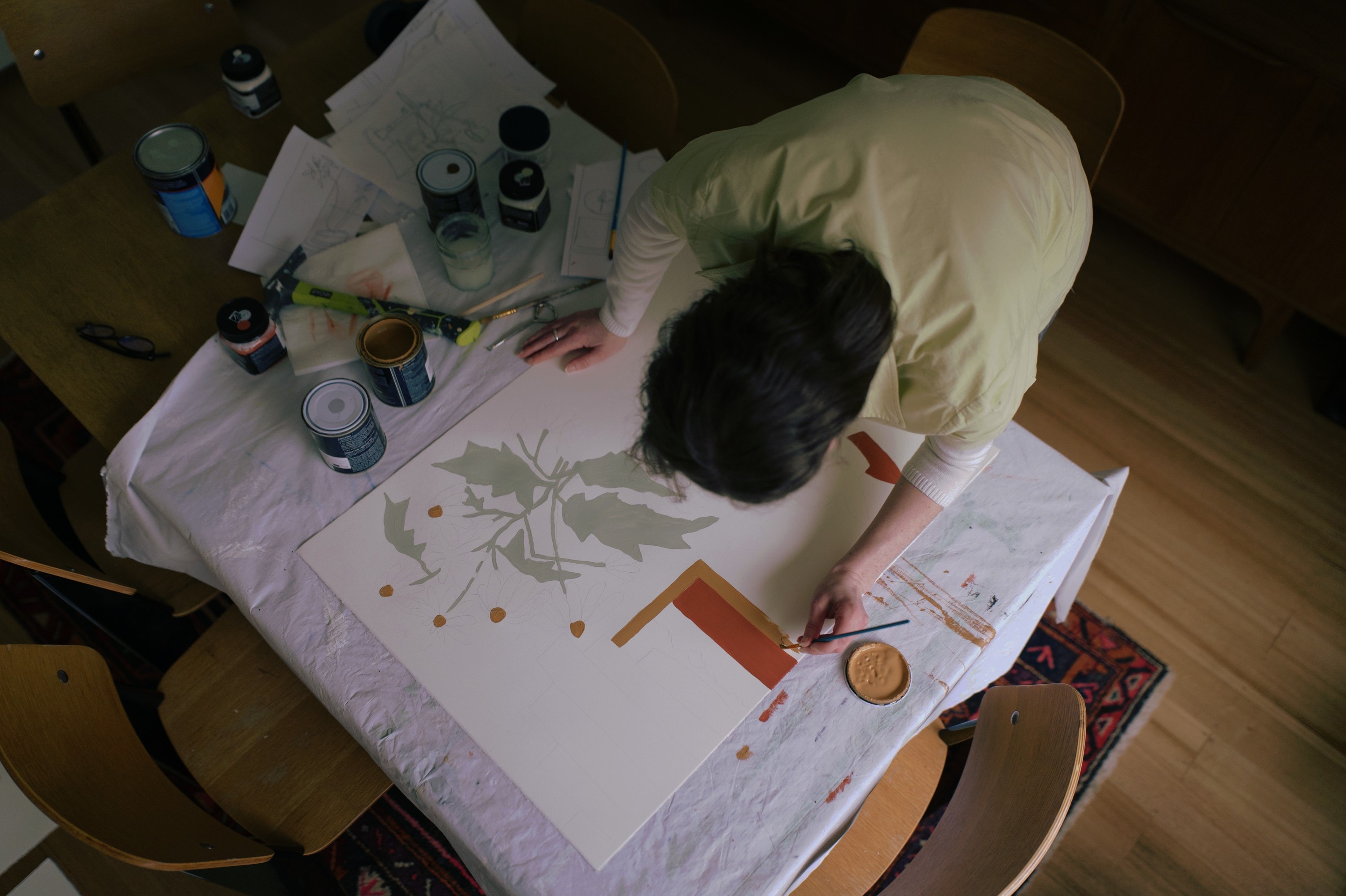 Angharad bent over her painting, seen from above, with paint tins around her