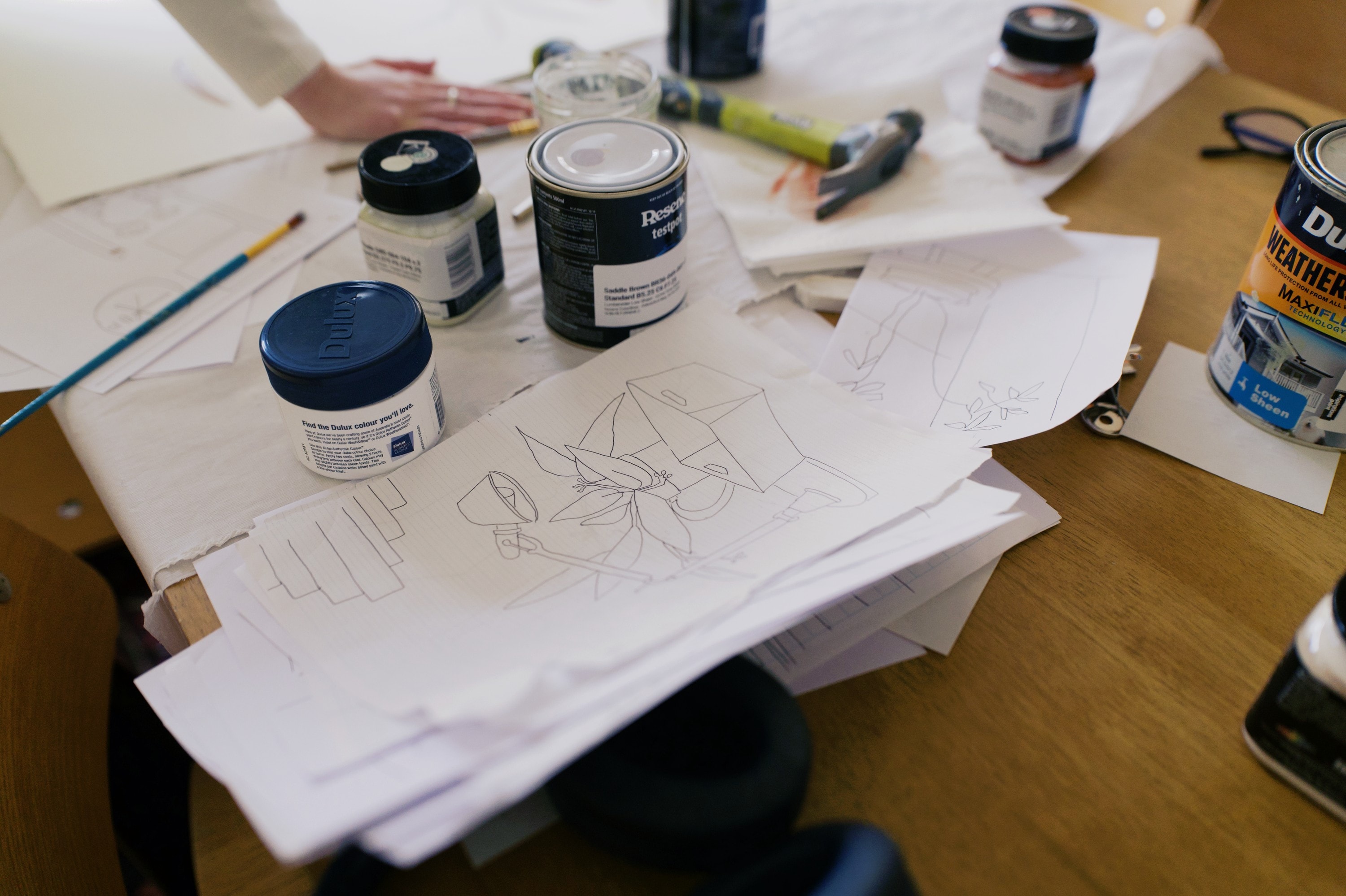 Pencil sketches on paper beside paint tins and brushes on the table