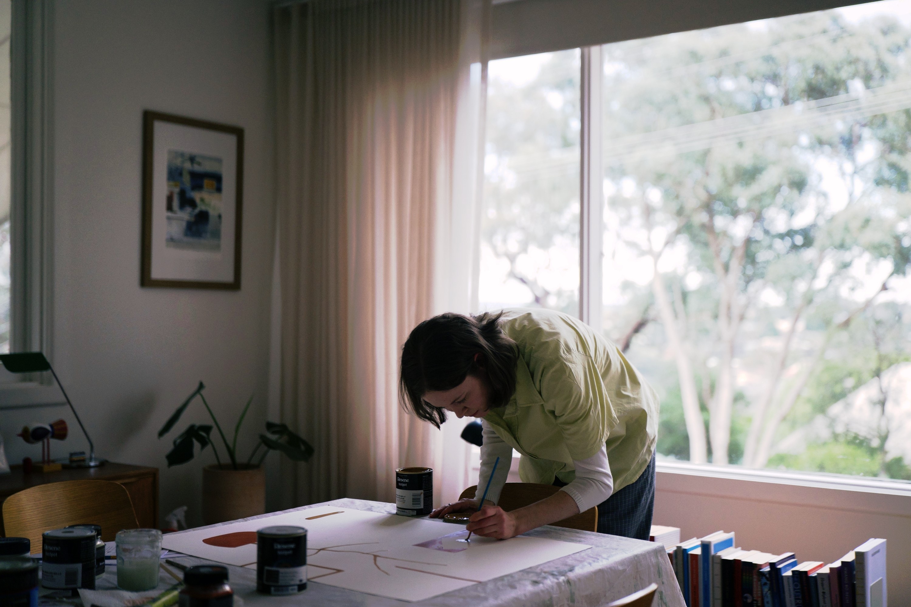 Angharad painting at the table in her sunroom, a framed print on the wall beside her