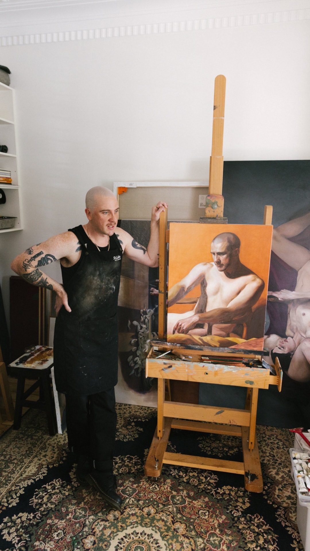 Anthony Stephens in a paint-flecked apron, leaning on his easel beside a portrait on an orange ground