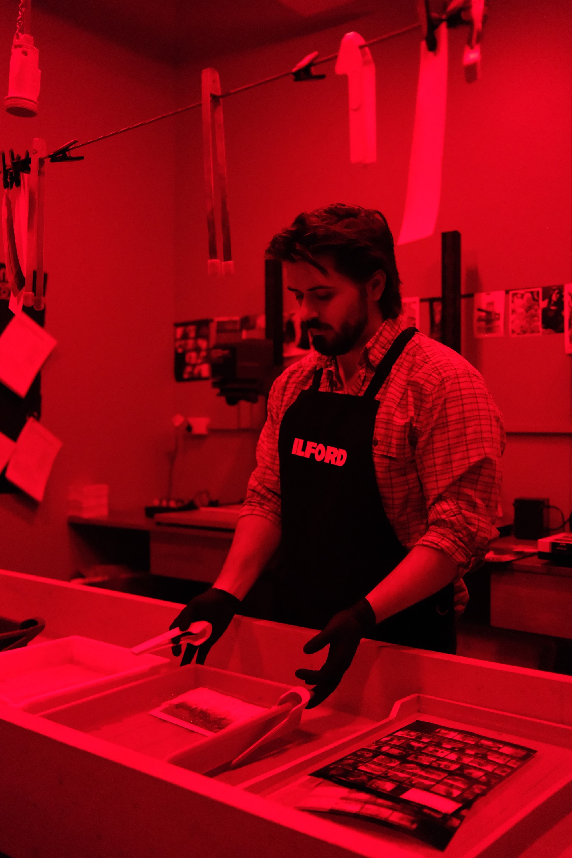 Dillon in gloves at the developing trays under the red safelight, a contact sheet in the last tray