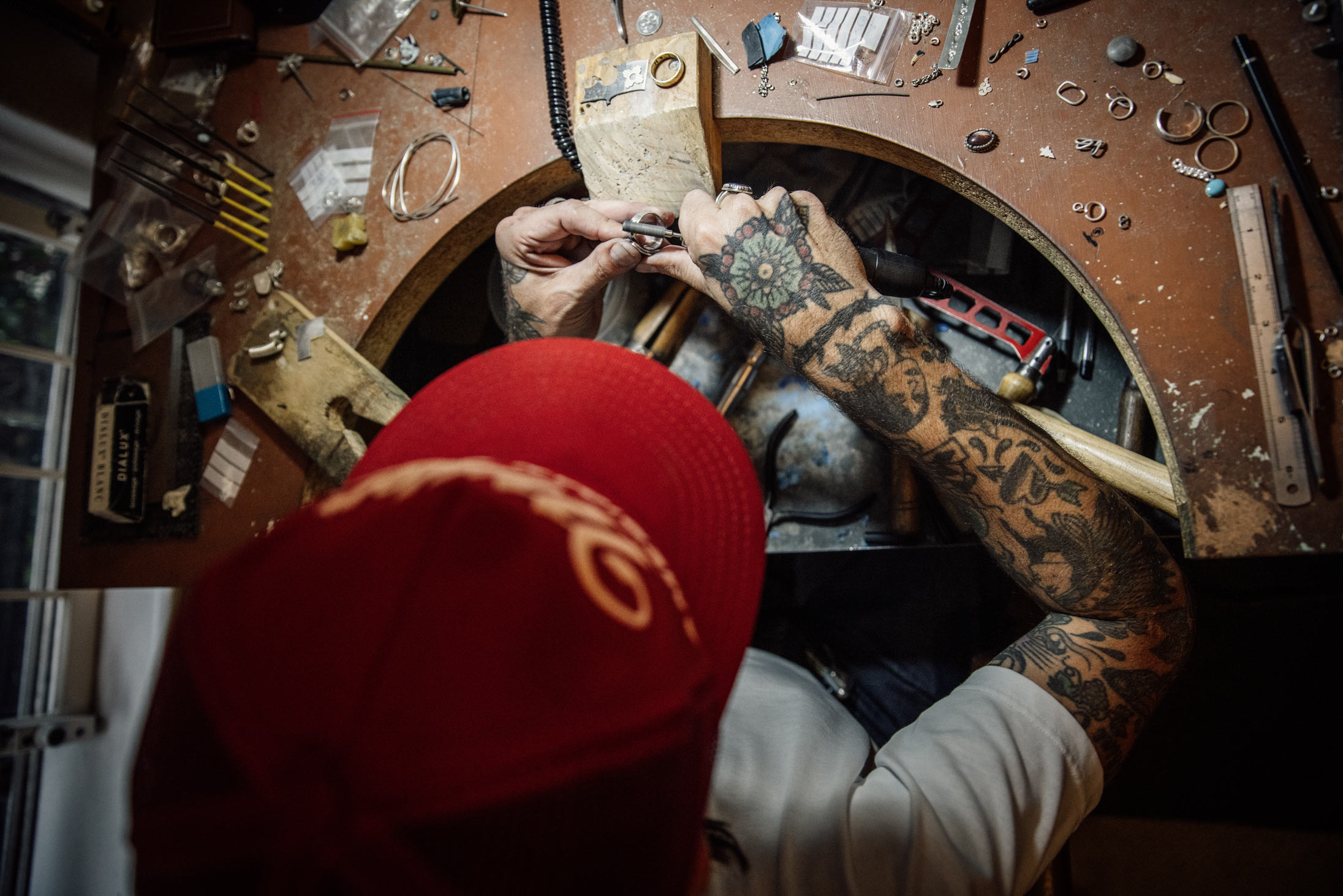 Adam at the bench from above, tattooed arms reaching into the bench pin over scattered silver and tools