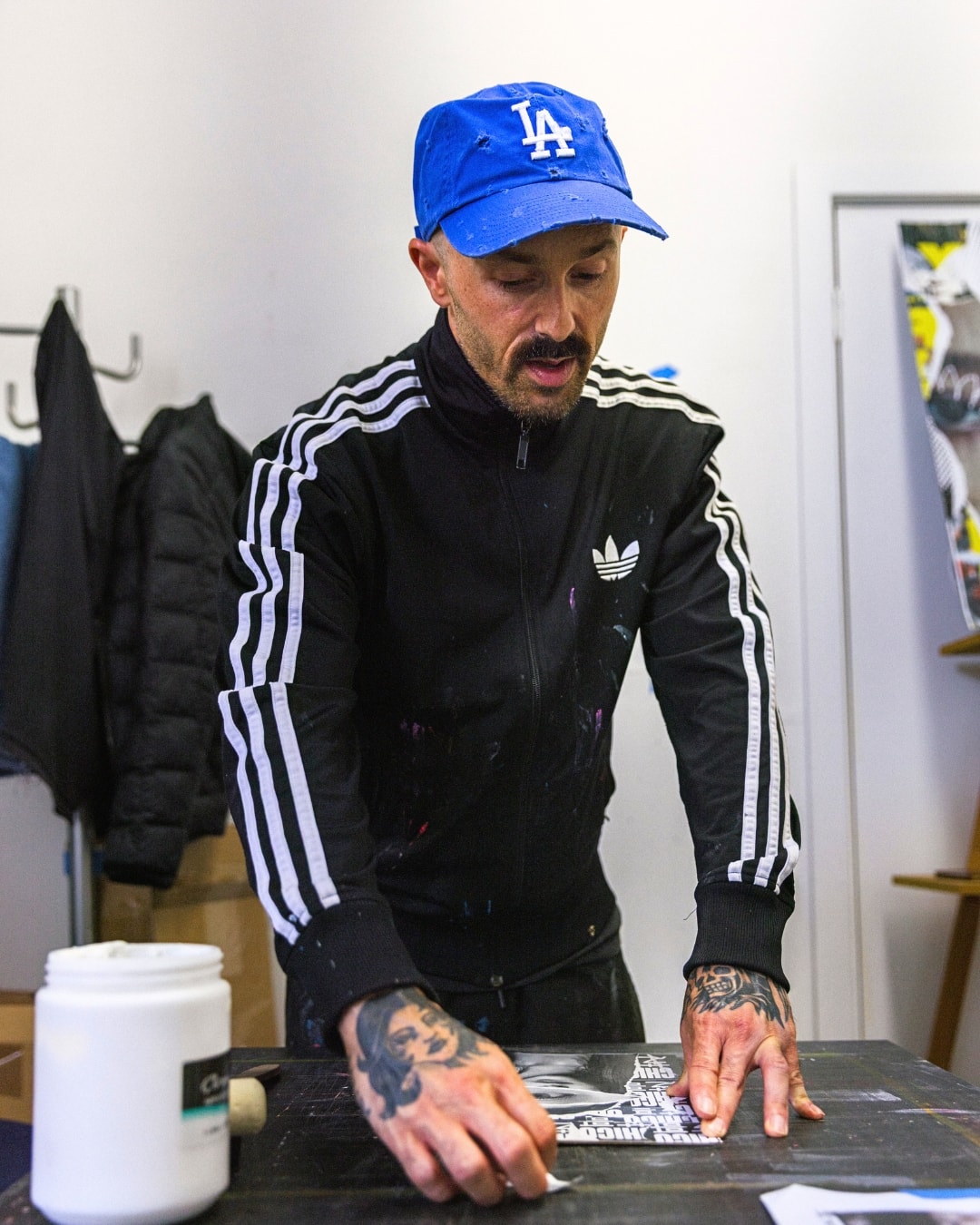Chico Leong in a blue LA cap leaning over his work table, smoothing down a collage