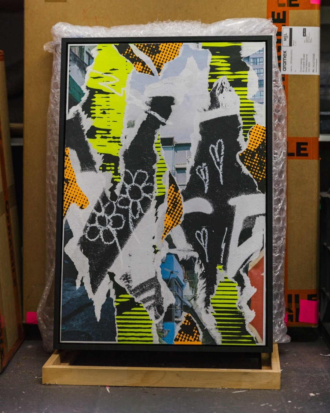A framed collage by Chico Leong in yellow, orange and black, propped on a stand in front of bubble wrap