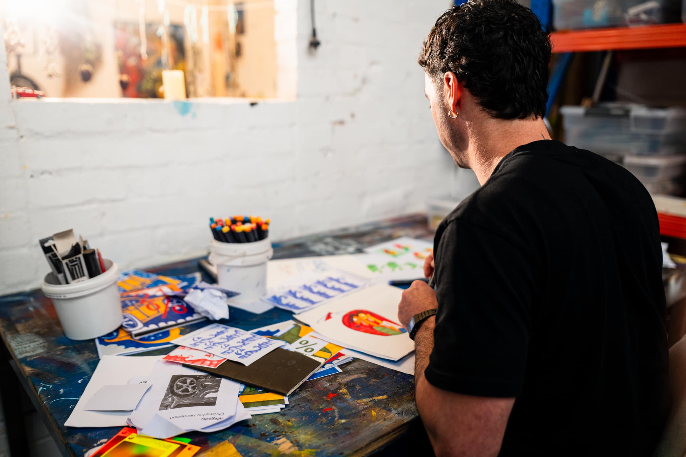 Over Chris’s shoulder at his desk, a tub of textas and sheets of drawings in front of him