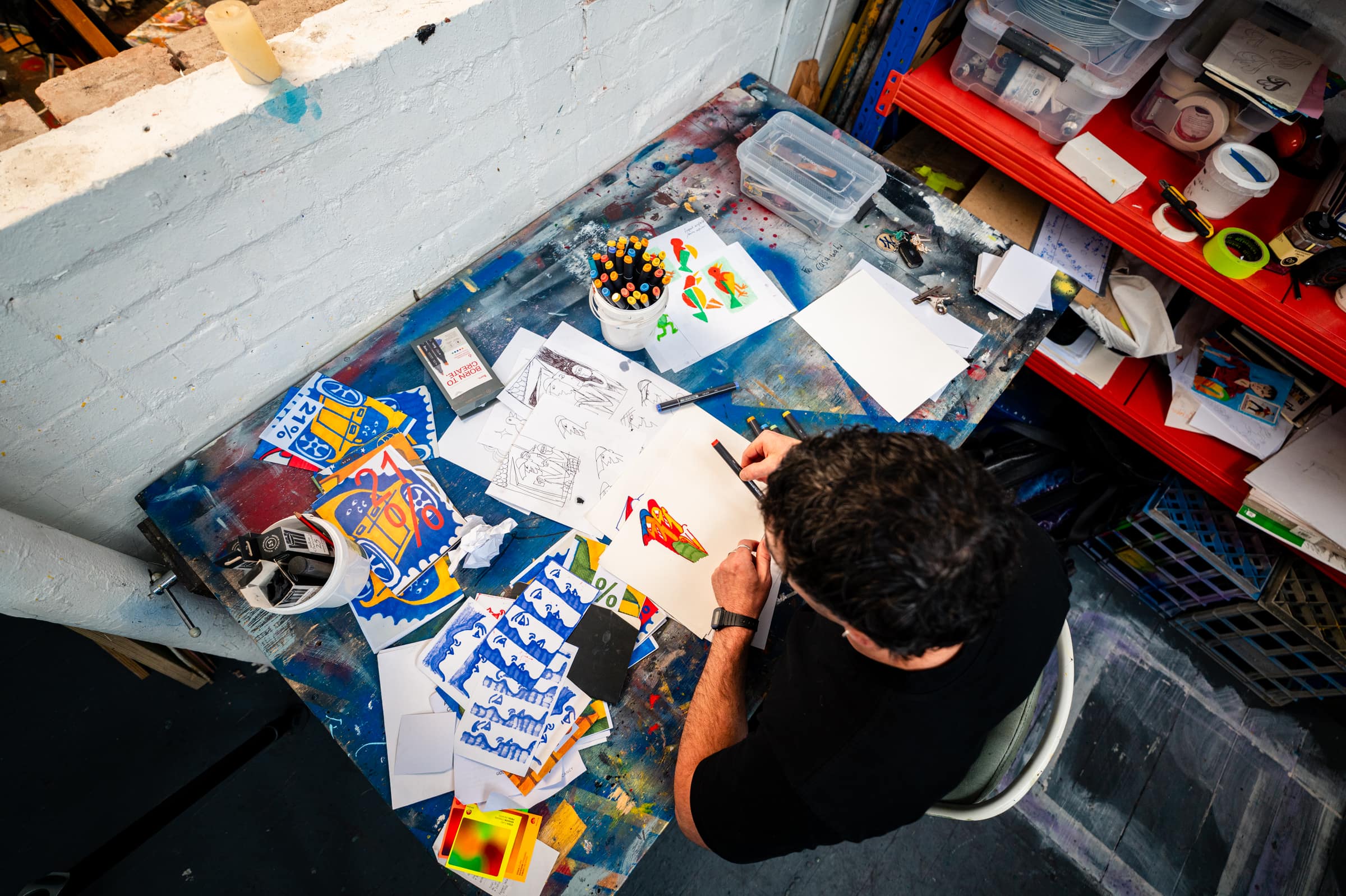 Looking down on Chris drawing at a desk covered in textas and blue Commodore drawings