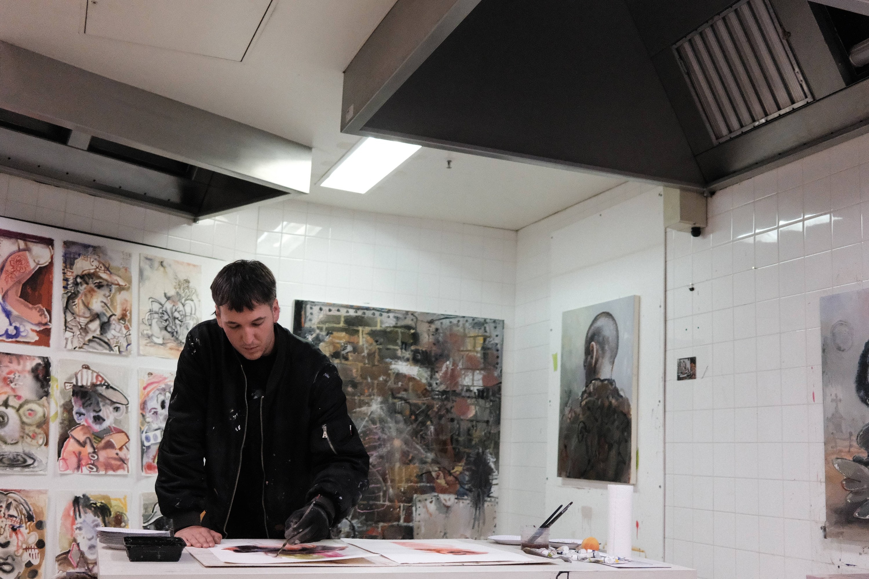 Rhys John Kaye painting on paper at a table in his tiled studio, works on paper pinned up behind him and a large abstract canvas on the wall