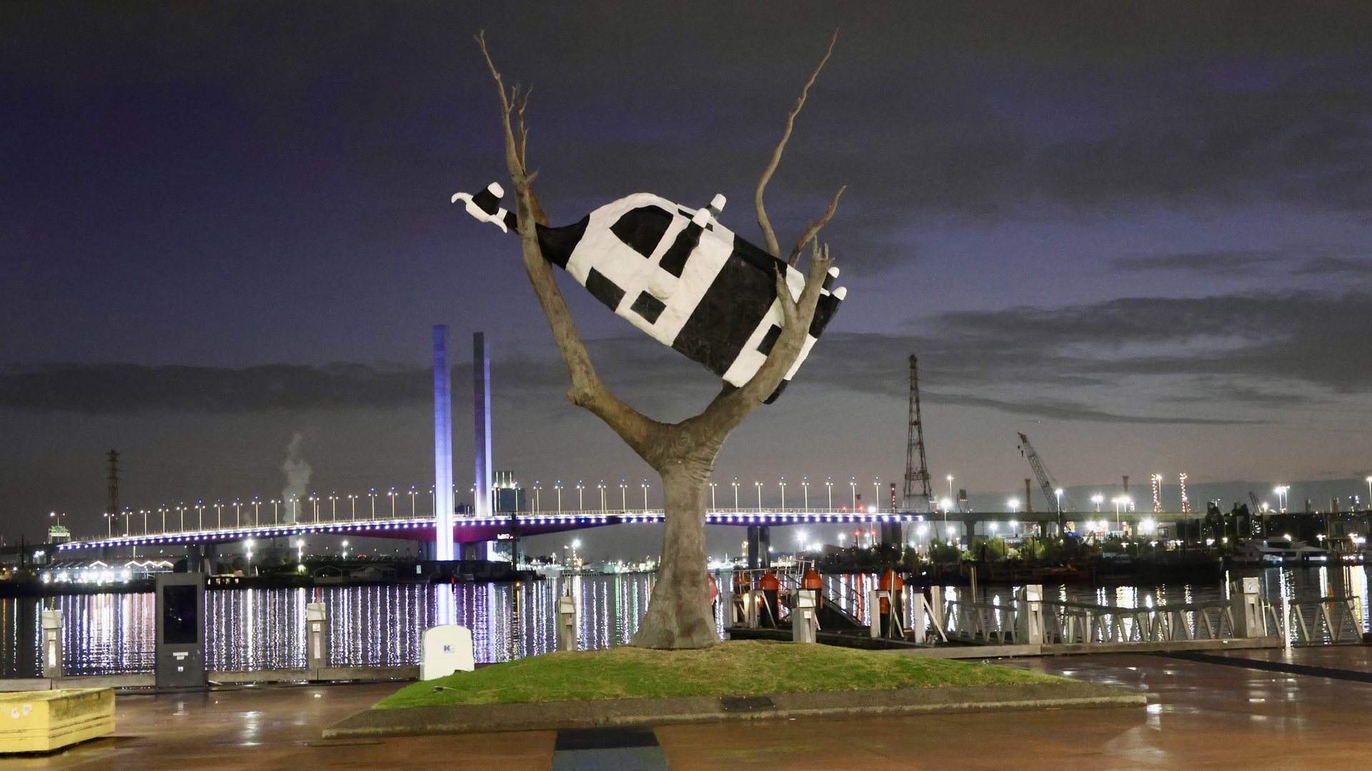 John Kelly's Cow Up a Tree, a black and white cow wedged in a bare tree, on the Docklands waterfront at dusk with the lit Bolte Bridge behind