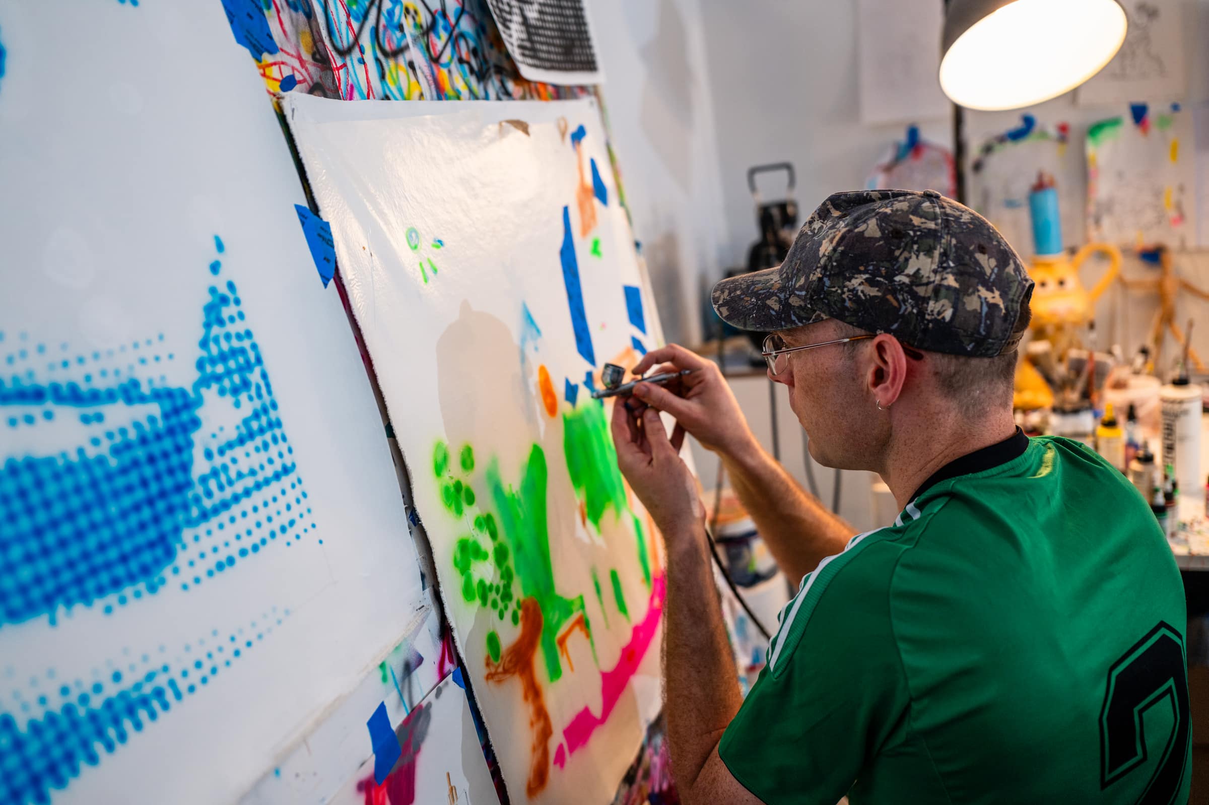 Liam airbrushing green shapes onto a sheet taped to the wall, a blue halftone painting beside him