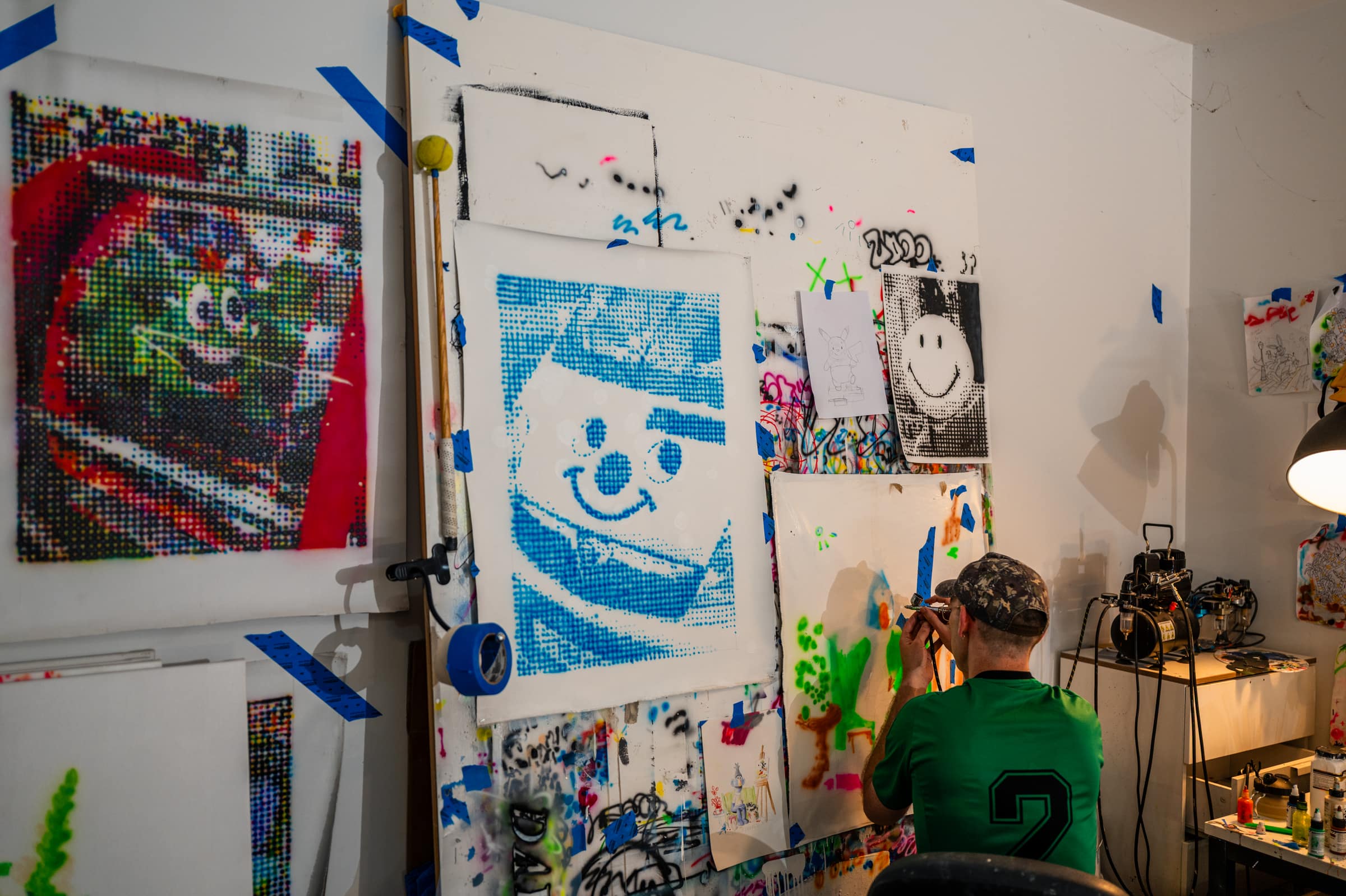 Liam airbrushing at his studio wall, beside a blue halftone clown face and a black and white smile