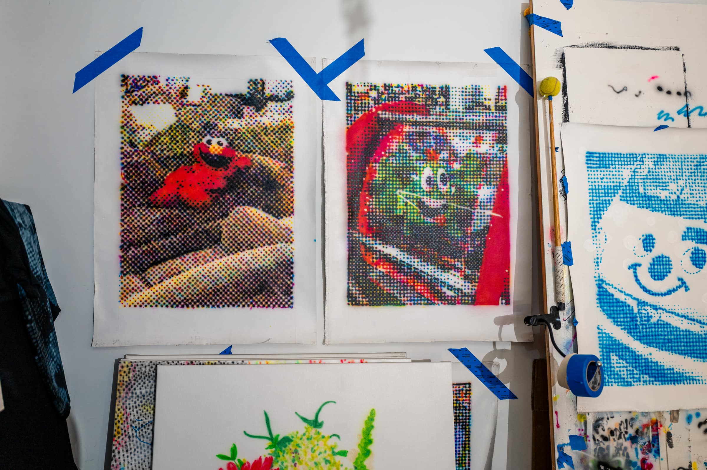 Halftone paintings taped to the studio wall: Elmo on a rock, a green character in red, and a blue clown face