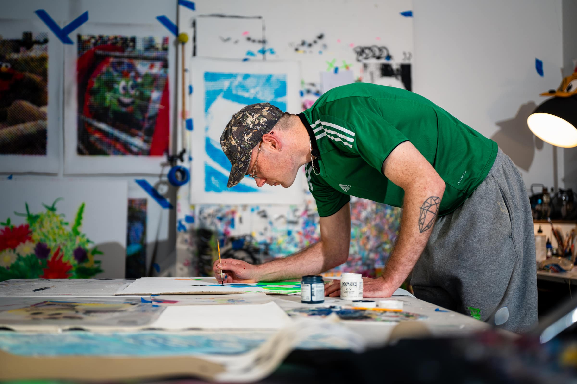 Liam leaning over his work table, painting a fluoro sheet with a fine brush, his halftone paintings on the wall behind