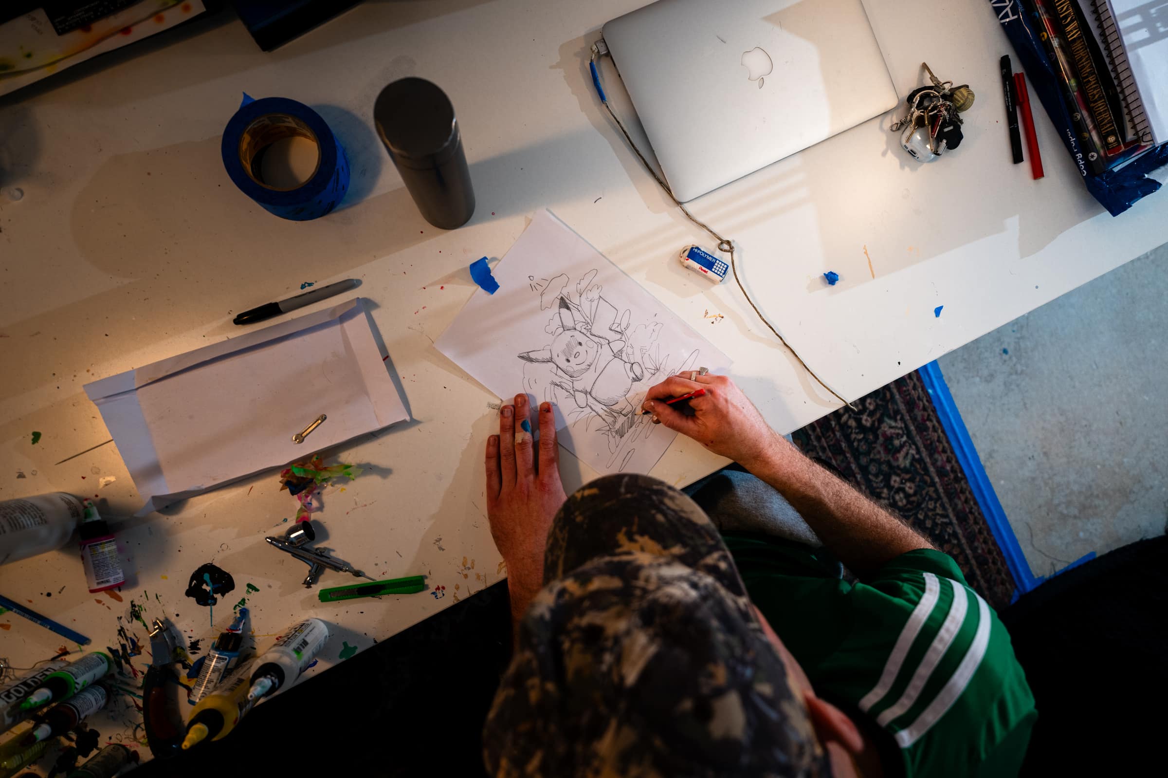Overhead view of Liam sketching a cartoon character in pencil at his desk, surrounded by tape, pens and a laptop