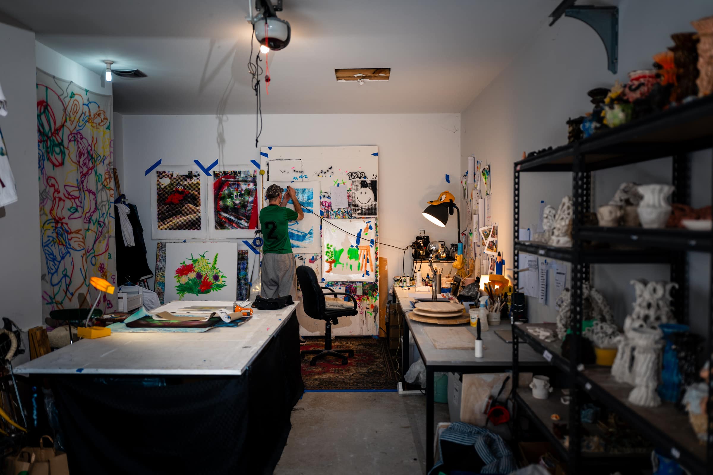 Liam's studio from the doorway: a long work table, shelves of toys and ceramics, and Liam at the far wall among his paintings