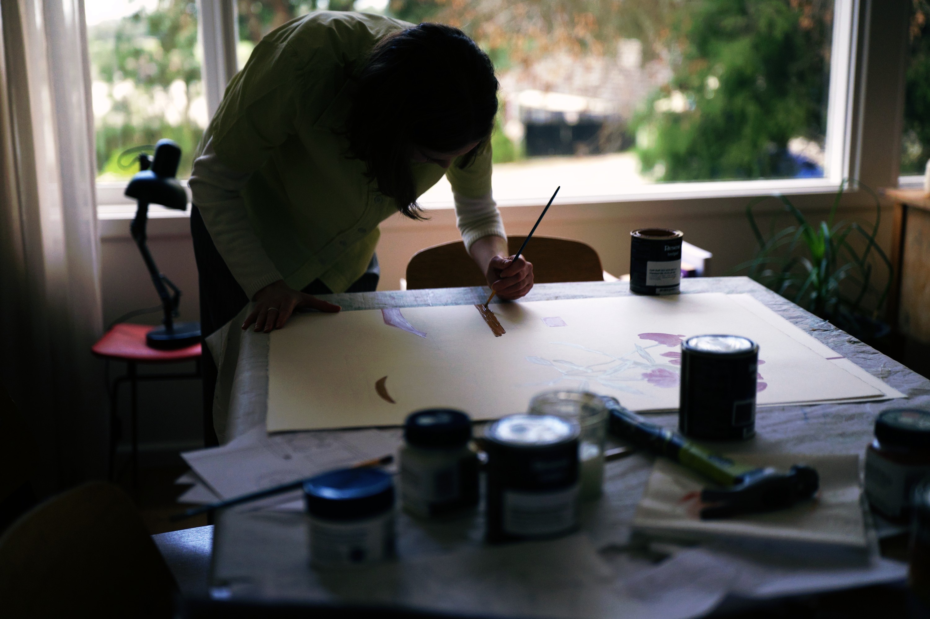 Angharad Neal-Williams leaning over a large sheet of paper at her table by the window, paint tins in the foreground