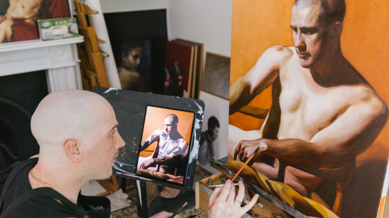 Anthony Stevens at his easel, glancing at a reference photo on a tablet beside a large portrait in oils on an orange ground