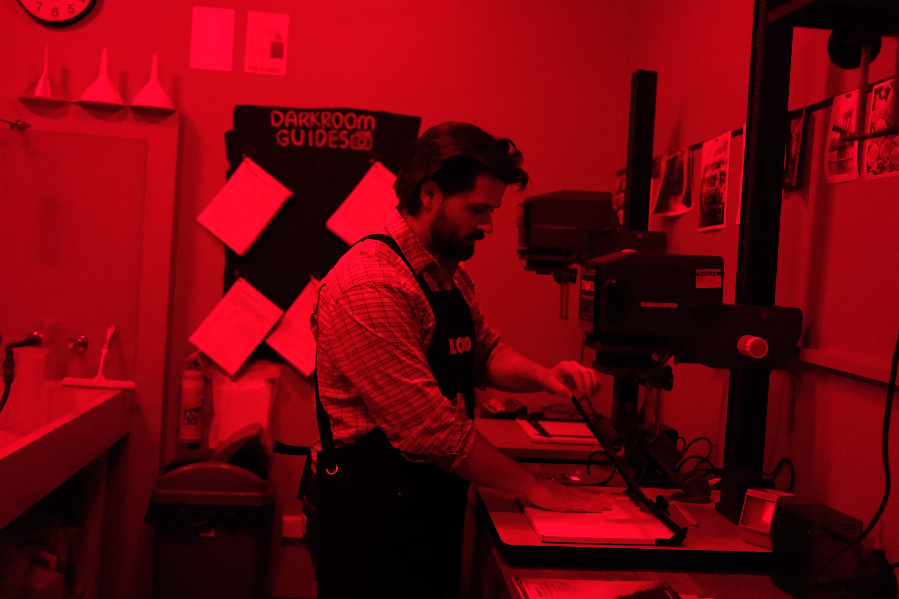 Dillon Jevon working under the red safelight at the enlarger in Brunswick Darkroom, with test strips pinned to the wall