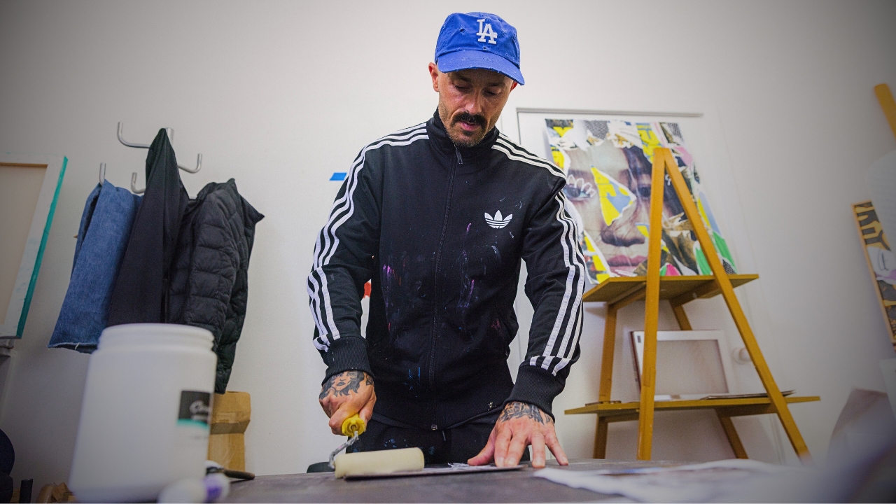 Chico Leong in a blue LA cap and black track jacket, rolling down a collage at his studio table