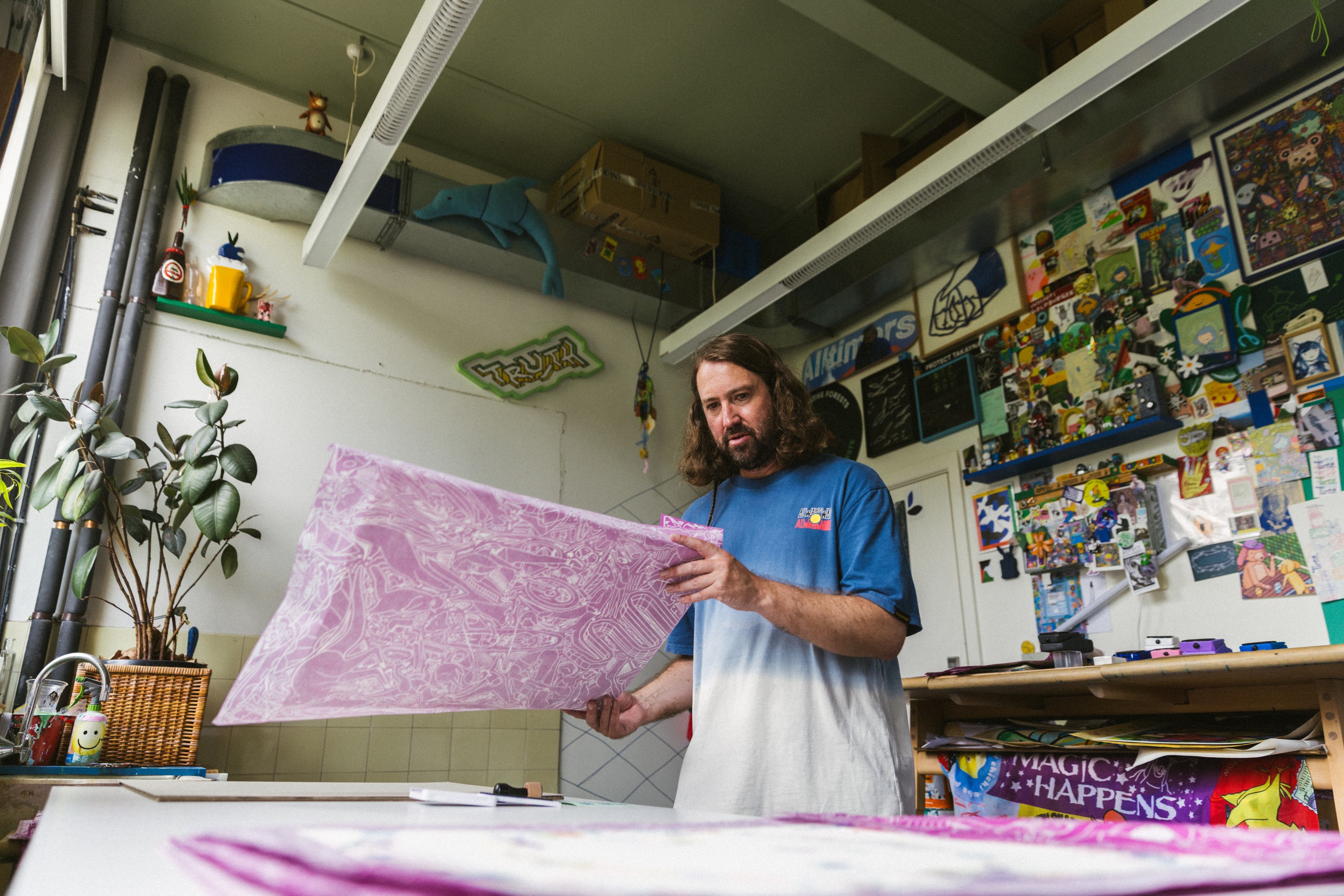David Booth holding up a large pink print in his studio, shelves of toys and drawings behind him