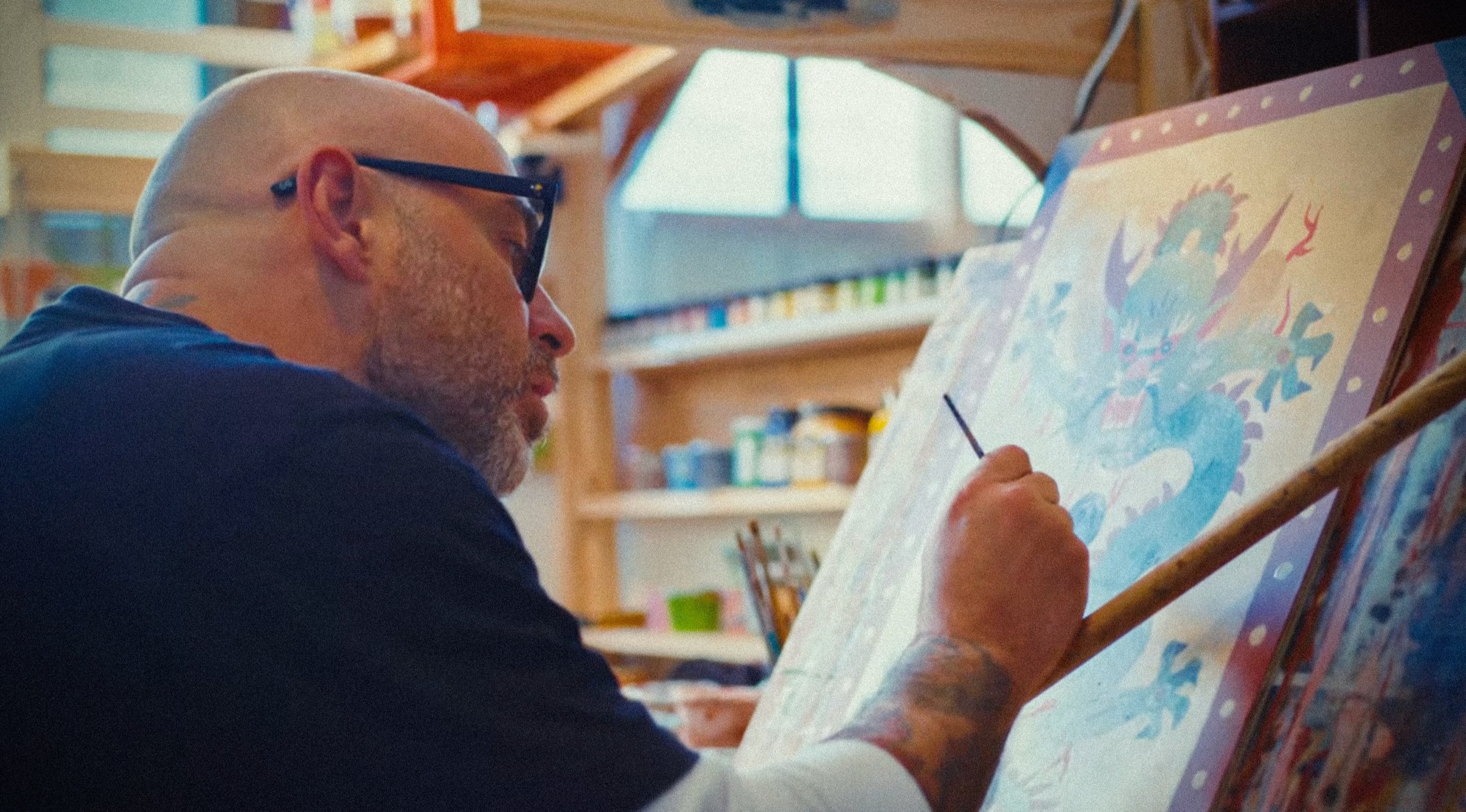 Jakob Morley at his easel painting a pale blue dragon inside a dotted lilac border, shelves of paint jars behind him