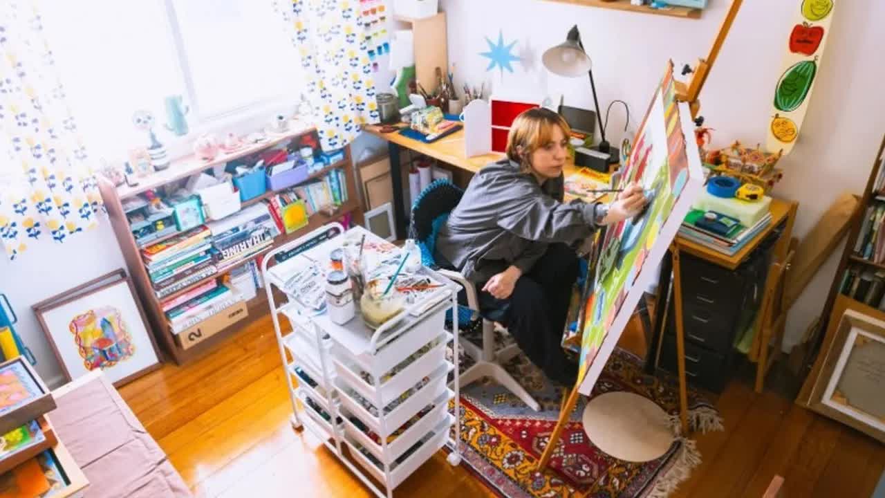 Melissa Grisancich painting a bright canvas on an easel in her studio, shelves of books and collected objects around her