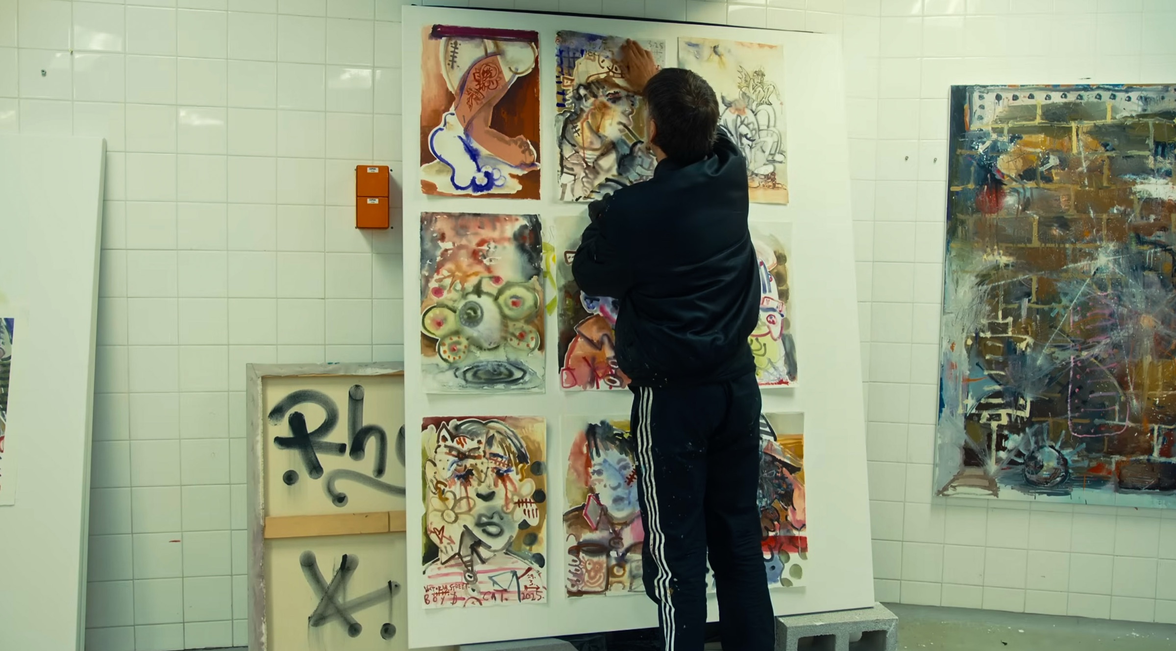 Rhys John Kaye from behind, pinning works on paper to a white board in his tiled studio, a large abstract canvas on the wall to the right