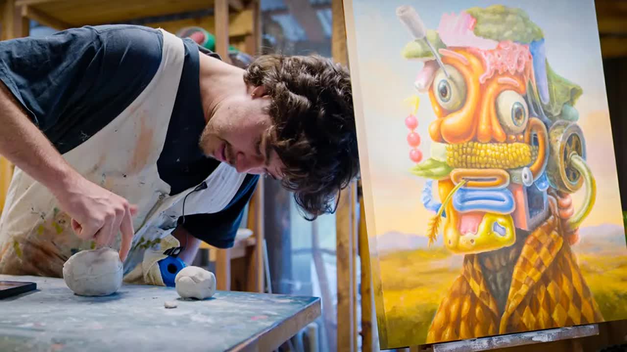 Sam Wilkinson leaning over a table shaping balls of clay, beside an oil painting of a bright, surreal character with a corn cob mouth